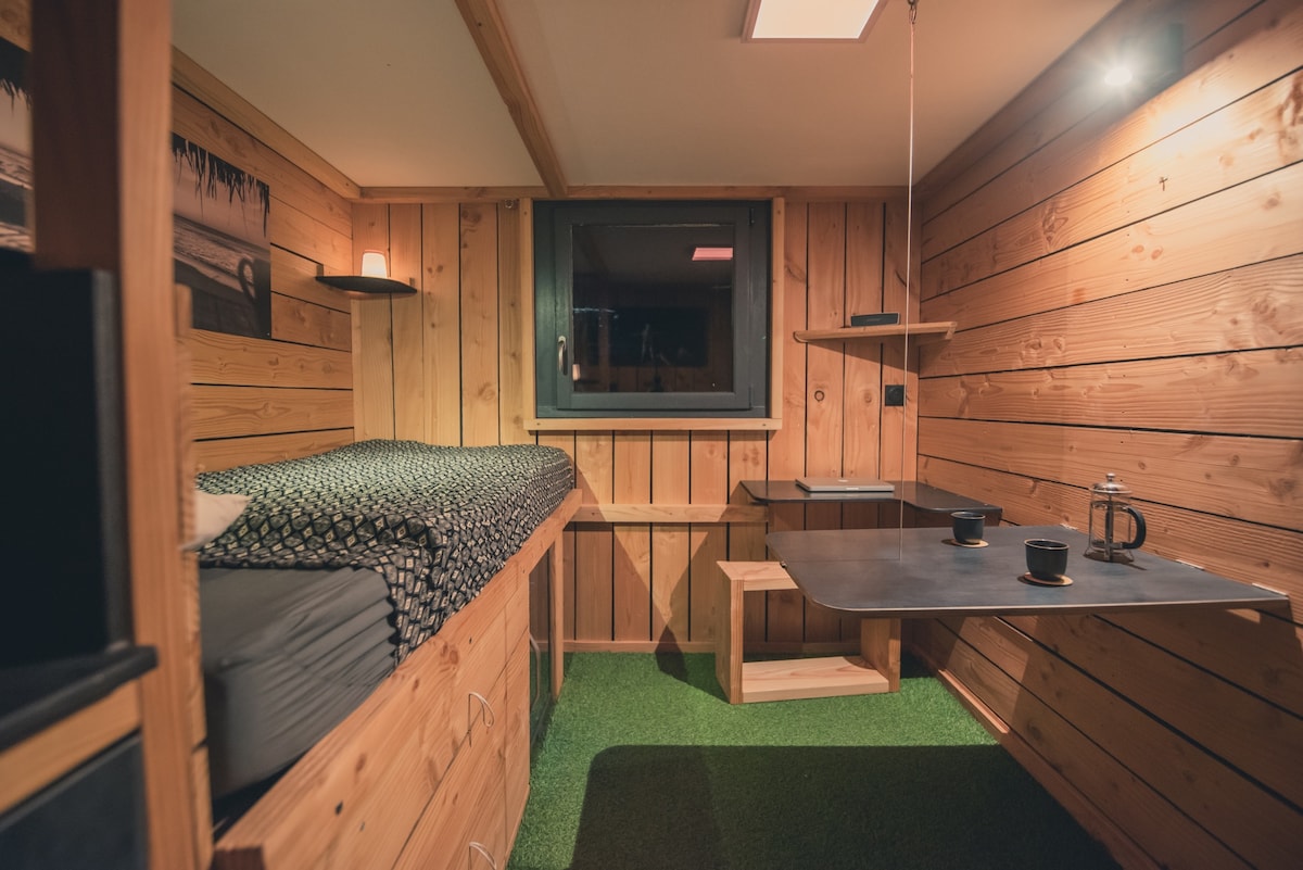 A cozy cabin room is surrounded by wood paneling, featuring a spacious bed with a patterned blanket. A simple work desk and a small table are positioned near a window, which offers natural light. Two cups sit atop the table, inviting relaxation.