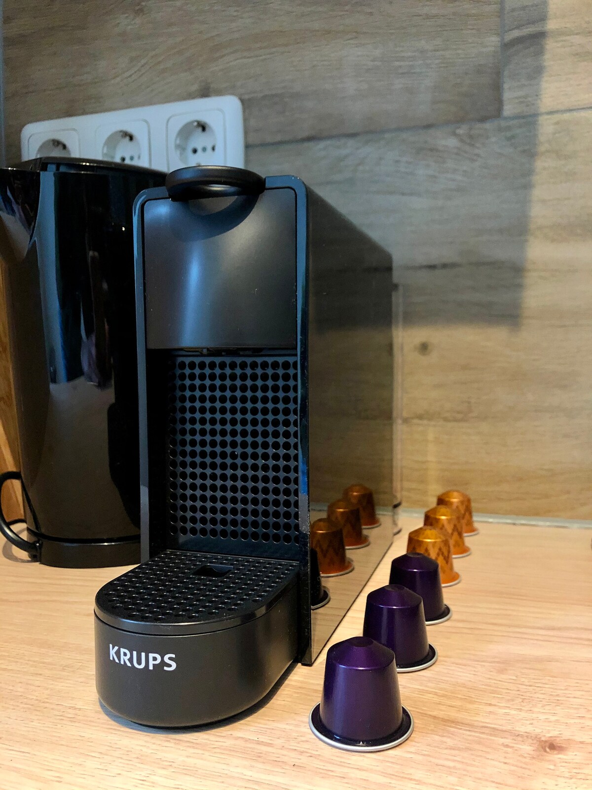 A coffee maker by Krups sits on a wooden counter, accompanied by various coffee capsules arranged neatly beside it. The sleek, modern design features a black finish, with a textured section for easy access to the coffee capsules.