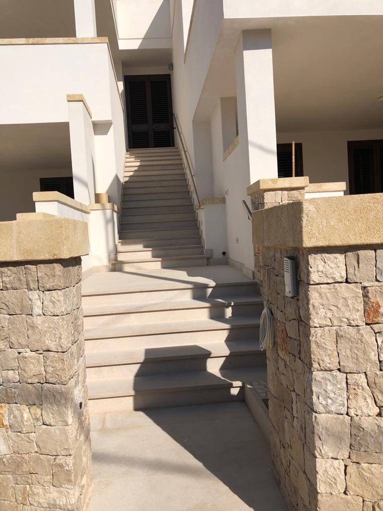 An exterior view captures a flight of stairs leading to a bright entrance, framed by stone pillars. The minimalist design emphasizes clean lines and natural light, while shadows enhance the stone texture. The area appears well-maintained and accessible.
