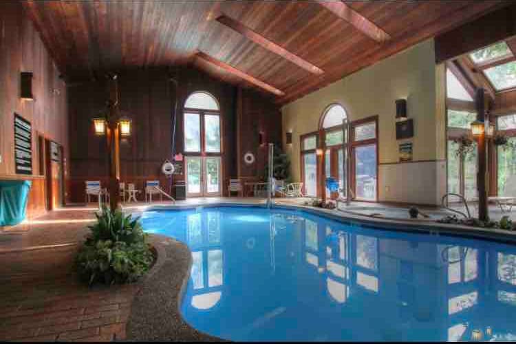 An indoor pool area features warm wooden beams above and large arched windows allowing natural light. The clear blue water is complemented by lush greenery at the edges and several lounge chairs positioned nearby, creating a tranquil space for relaxation and leisure.