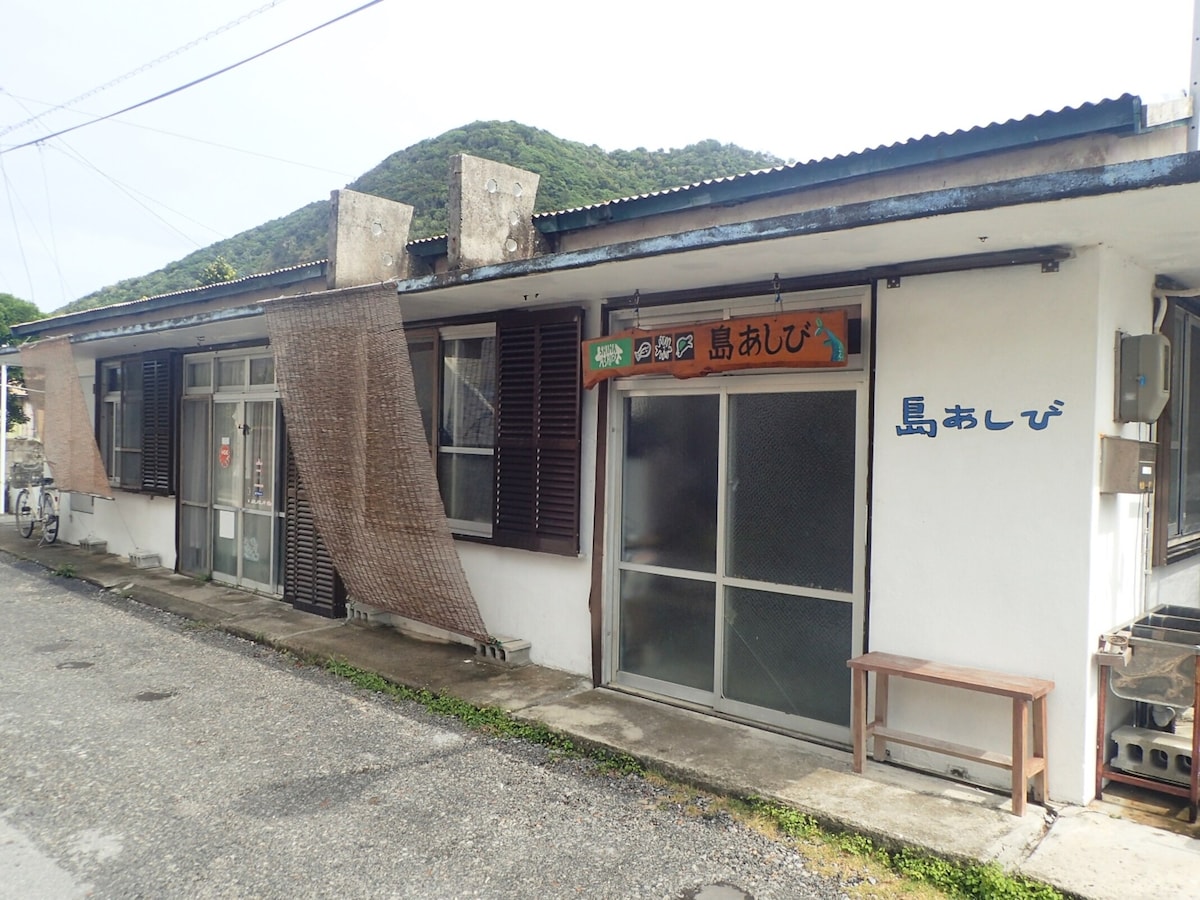 The exterior of a single-story building is shown, featuring a simple design with white walls and dark wooden shutters. A sign reading '島あしび' is visible above the entrance. The surrounding environment includes natural greenery and a gravel pathway leading to the entrance.