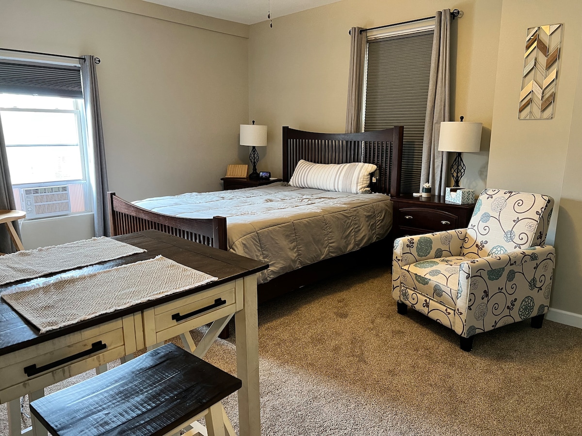 The bedroom features a spacious bed with light-colored bedding, flanked by two bedside tables, each with a lamp. A small dining table sits near the window, accompanied by a decorative chair. Natural light enters through the window, enhancing the neutral tones of the room.