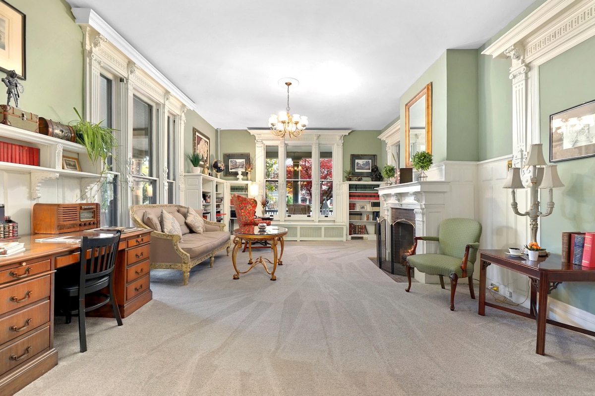 A spacious common area is depicted, featuring large windows that invite natural light. Comfortable seating is arranged near a classic fireplace, with a wooden desk and chair positioned nearby. Bookshelves line the walls, filled with books and decorative items, enhancing the charm of the room.