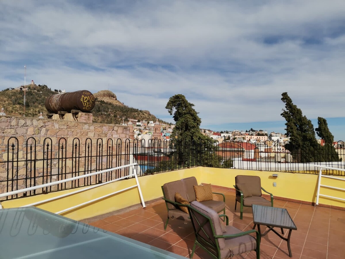 An open terrace is featured, surrounded by a low black railing, offering views of a cityscape with colorful rooftops and distant hills. Two brown chairs and a small table are present, inviting relaxation in a sunny outdoor setting.
