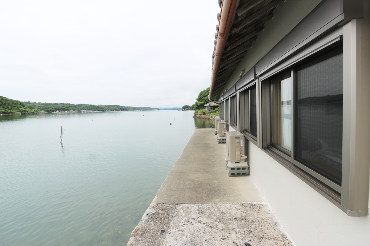 A serene outdoor space borders the calm water, featuring a wide walkway along the shoreline. Several air conditioning units are positioned beside large windows that offer views of the water and surrounding greenery.