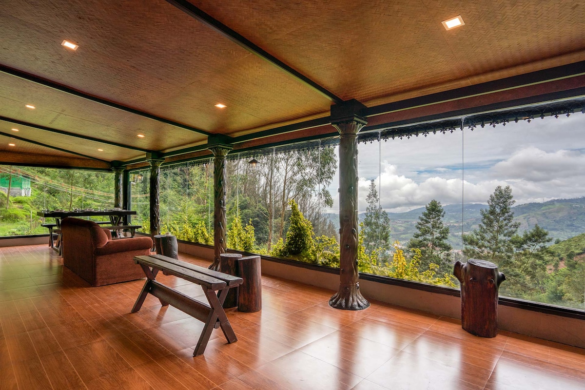 A spacious common area features floor-to-ceiling glass windows, offering expansive views of the surrounding landscape. Natural wood accents can be seen in the furniture, including a long bench and a table. Soft lighting enhances the serene atmosphere, inviting relaxation and connection with nature.