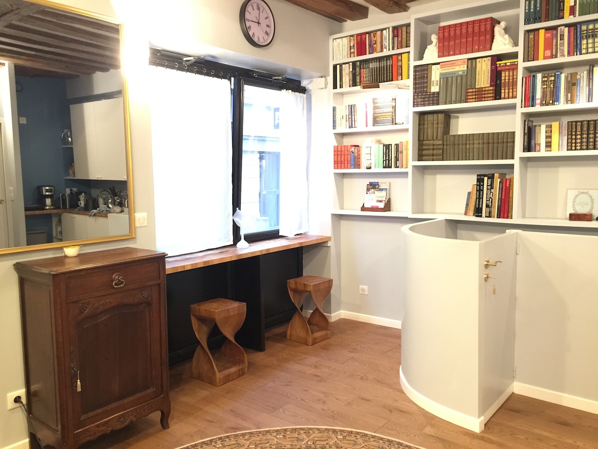 A cozy reading nook features a wooden shelf filled with books and a large window allowing natural light to enter. Two wooden stools provide seating, complemented by a decorative mirror on the wall. The warm wooden beams overhead add character to the space.