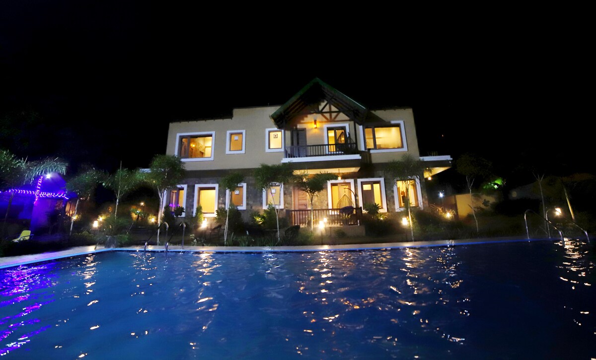 An illuminated villa is viewed from a swimming pool at night, showcasing multiple rooms with warm lights glowing from the windows. Lush greenery surrounds the property, enhancing the tranquil ambiance, while water reflects vibrant colors from the surrounding lighting.