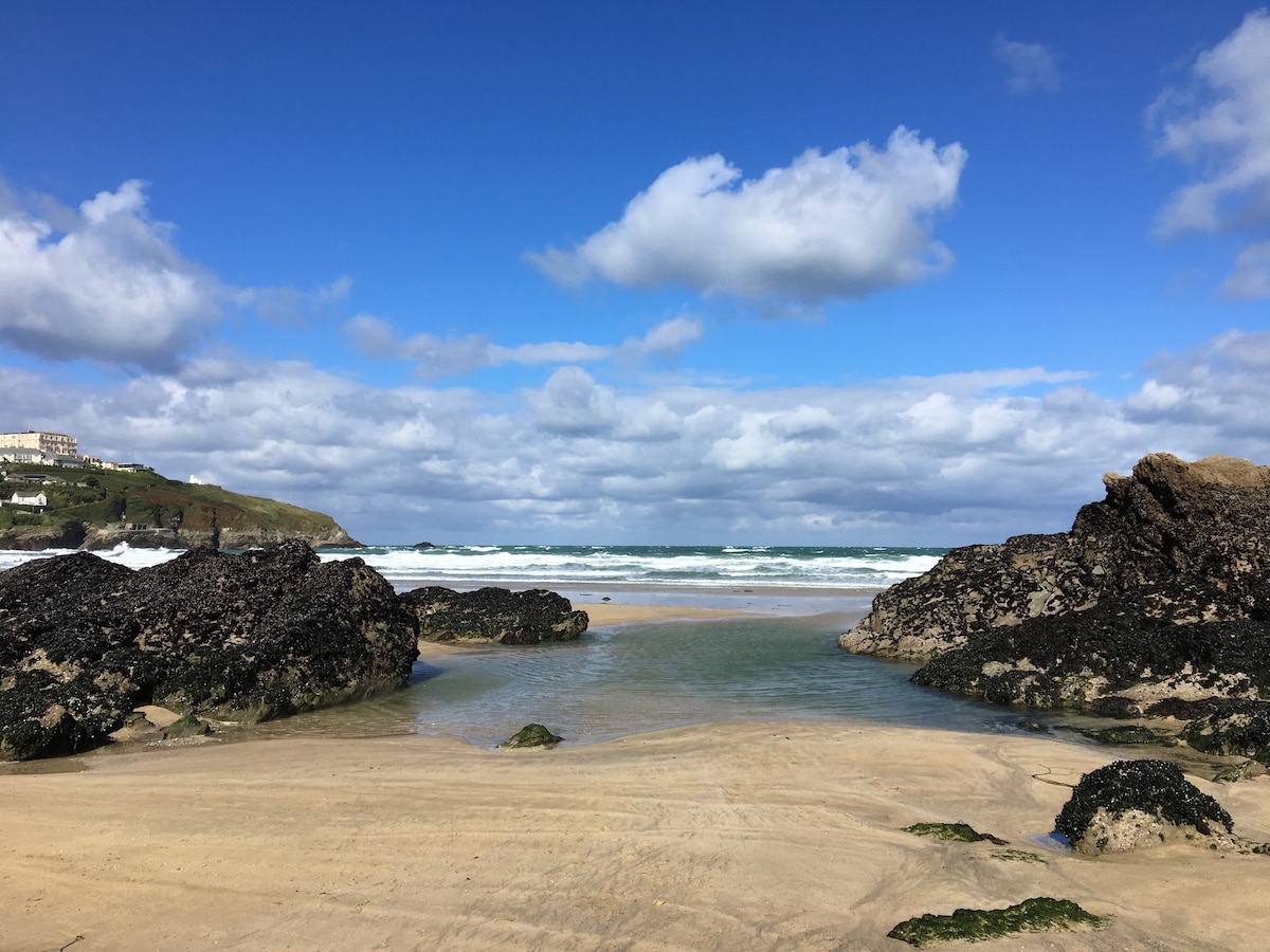 A serene beach scene is captured, showcasing golden sand bordered by rugged rocks. Gentle waves roll towards the shore under a bright blue sky accented with fluffy white clouds. The calm waters reflect the clear sky, creating a tranquil coastal atmosphere.
