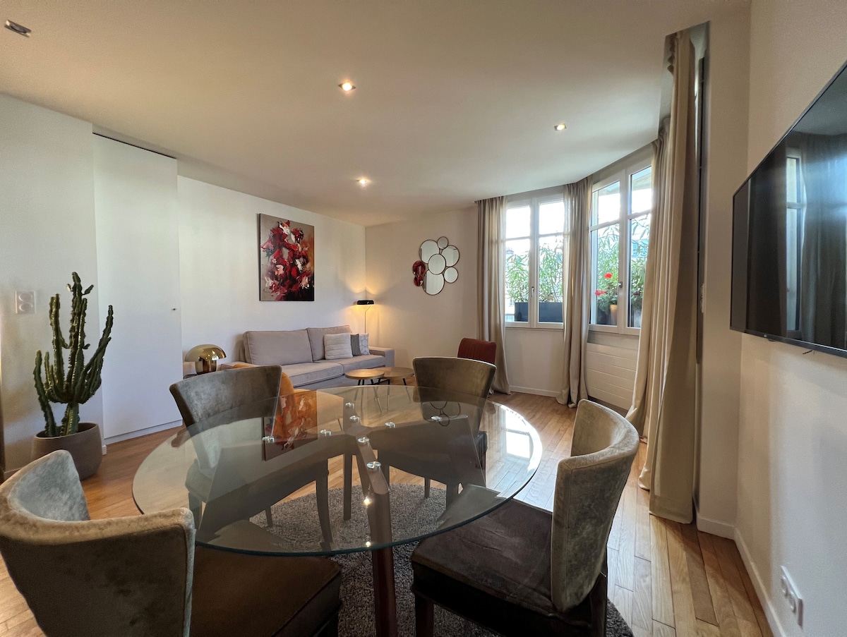 A circular glass table is surrounded by four upholstered chairs, offering a seating area. A streamlined sofa is positioned against the wall, accompanied by a wall-mounted TV. Large windows with potted plants provide natural light, while a decorative wall piece adds subtle character.