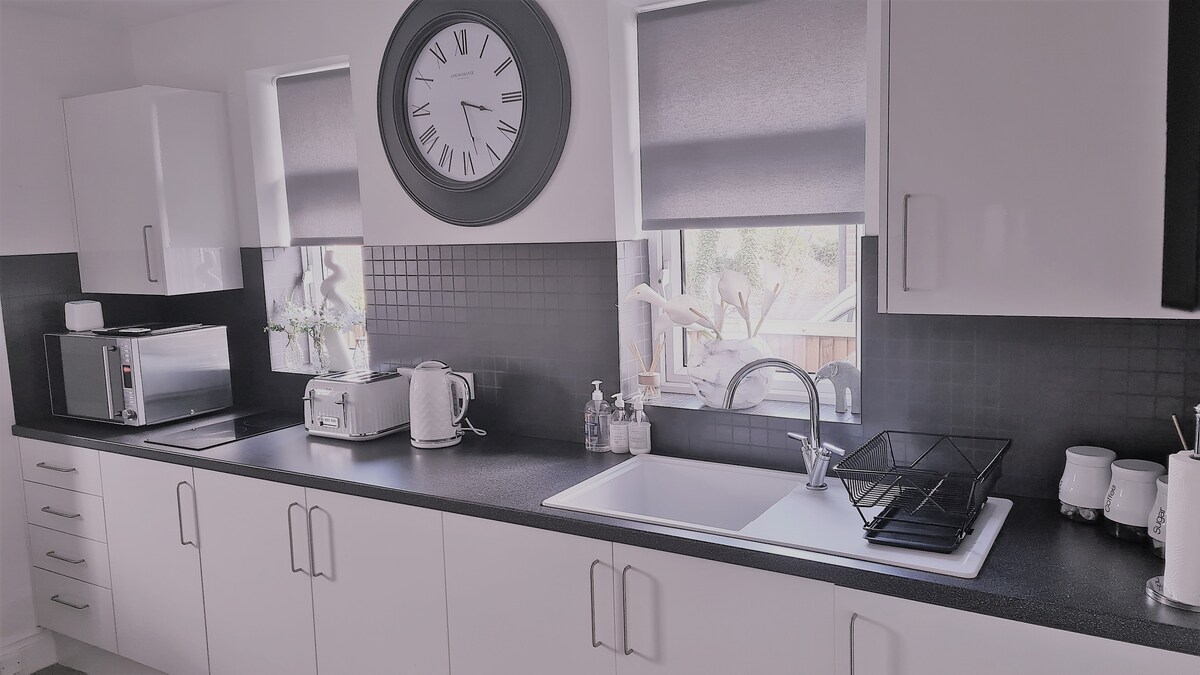 A modern kitchen features sleek white cabinetry and a dark countertop. Natural light streams in through two windows, adorned with roller shades. A clock is mounted on the wall, complementing the stainless steel appliances, including a microwave and kettle. Kitchen utensils are neatly arranged beside the sink.