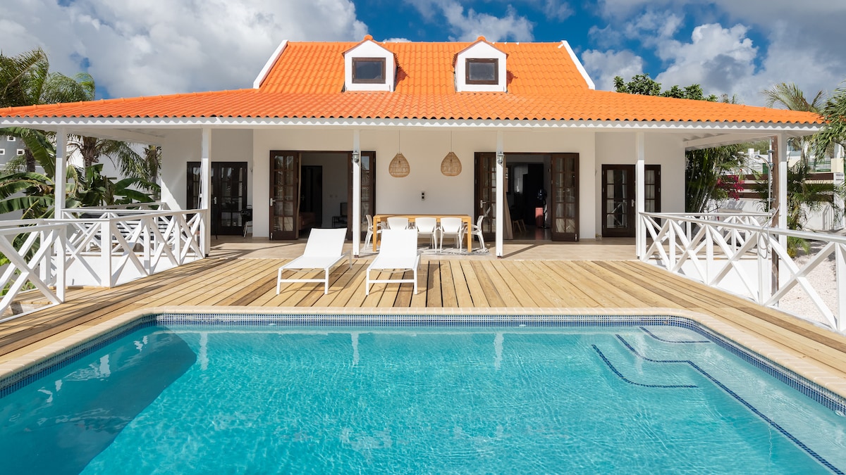 The exterior of the villa features a vibrant orange roof and spacious wooden deck. Lounge chairs are positioned beside a refreshing pool, inviting relaxation. The covered patio area includes a dining table and is framed by palm trees, enhancing the tropical ambiance.