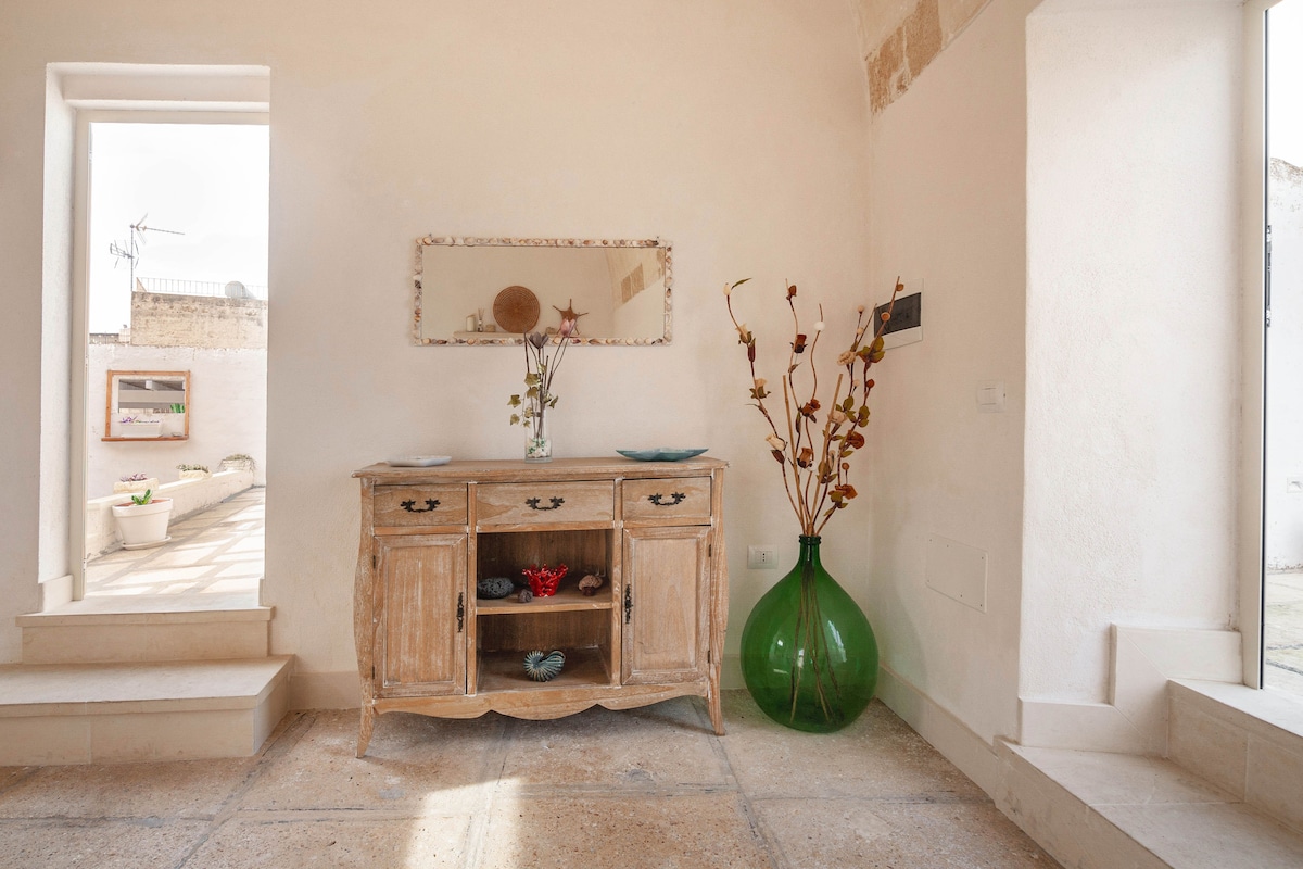 An entrance area features a rustic wooden sideboard, complemented by a decorative mirror and a vibrant green vase filled with dried flowers. Soft natural light illuminates the space through windows on either side, enhancing the warm and welcoming environment.