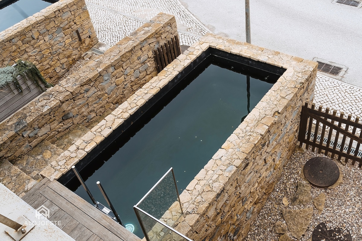 A private pool is outlined by rustic stone walls, providing a tranquil escape in the outdoor area. The pool's still water reflects its surroundings and is accessible via a clear glass door. The pool area is set against a quaint cobblestone backdrop.
