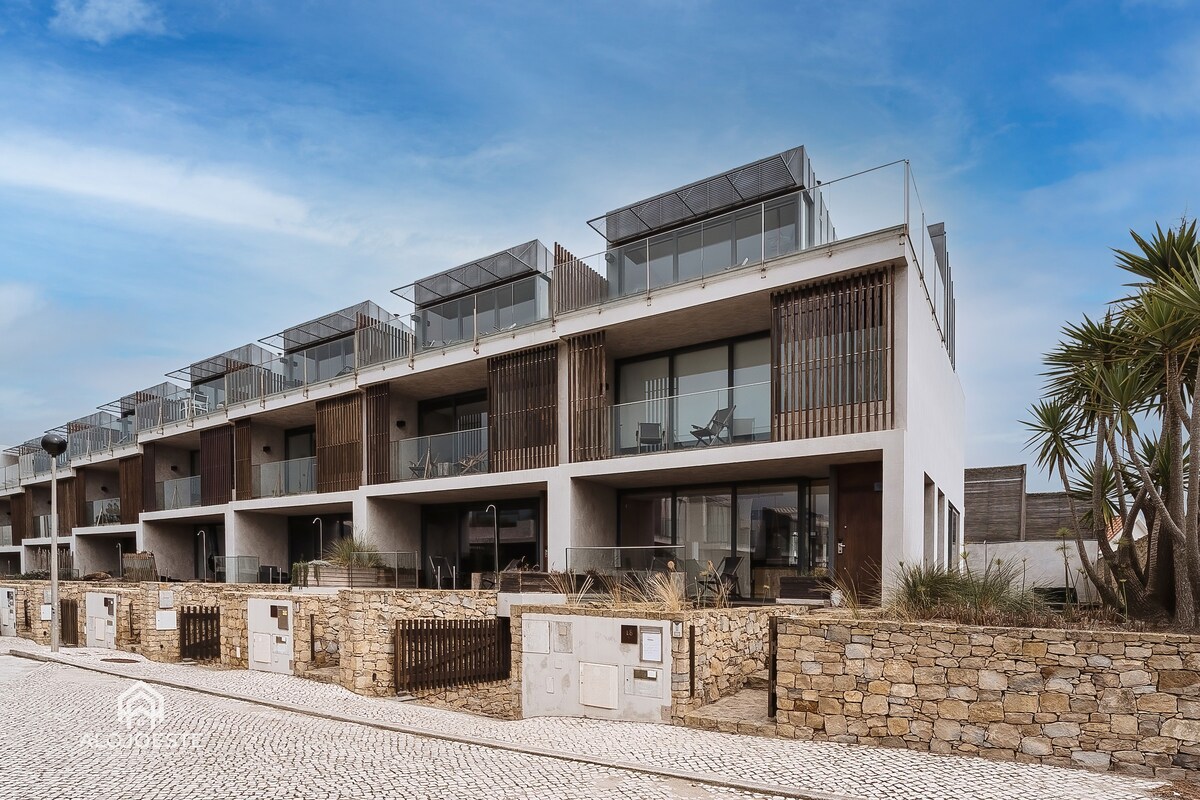 A row of modern villas features balcony terraces with glass railings, blending stylish architecture with natural elements. Each unit is framed by palm trees and a stone boundary wall, creating a welcoming exterior that complements the coastal environment.