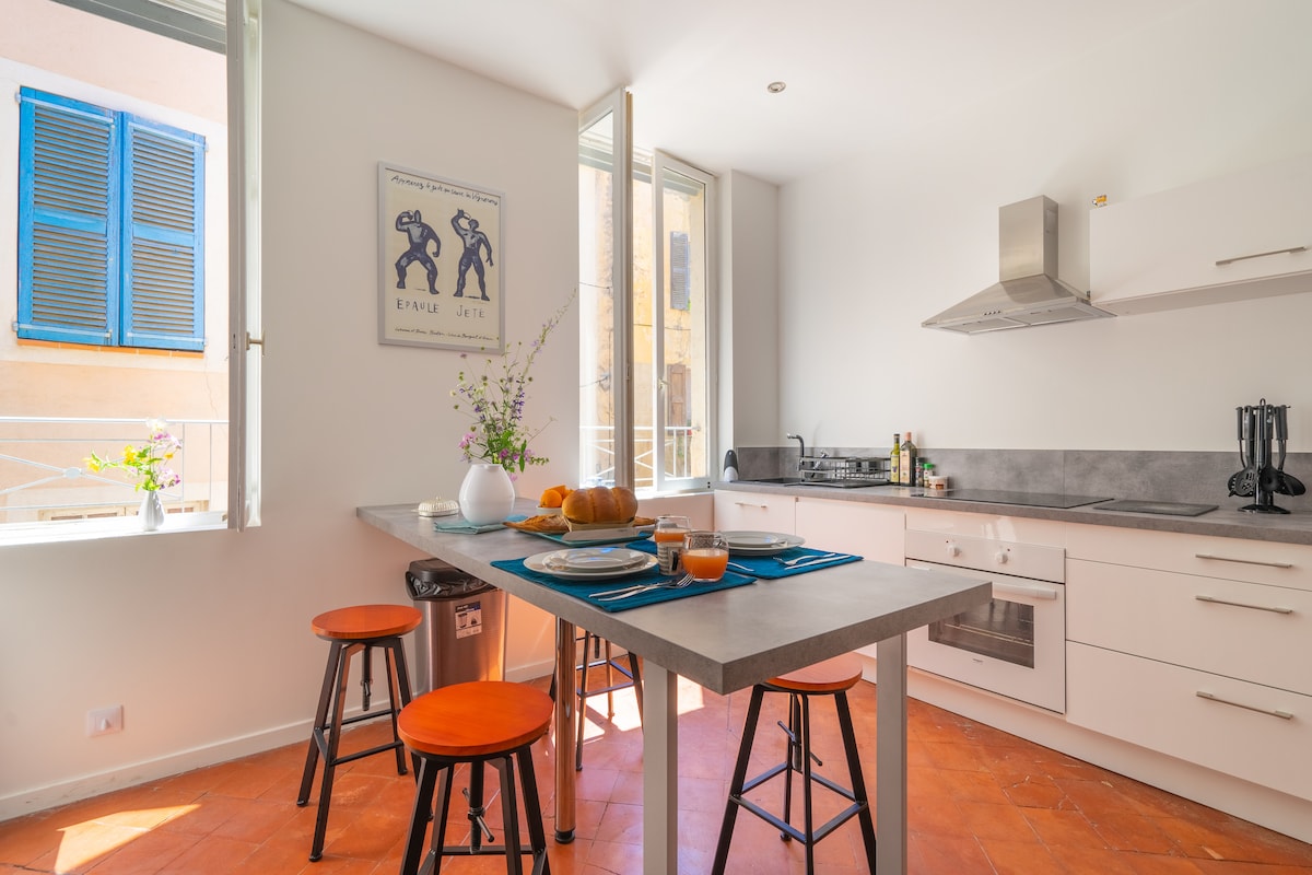 A modern kitchen is highlighted by a central table, adorned with colorful tableware and a fruit display. Large windows allow natural light to fill the space, complemented by wooden stools with orange upholstery. Sleek appliances and a minimalist design enhance the area.
