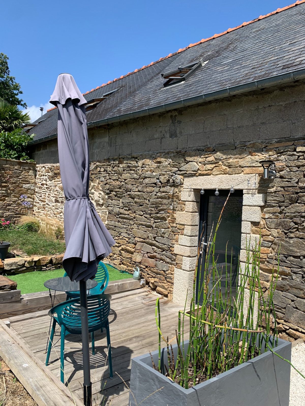 A charming stone wall provides a rustic backdrop for a sunlit terrace. A tall parasol stands beside modern teal chairs, while a planter with green plants adds a touch of nature to the area. Clear skies enhance the spacious outdoor setting.