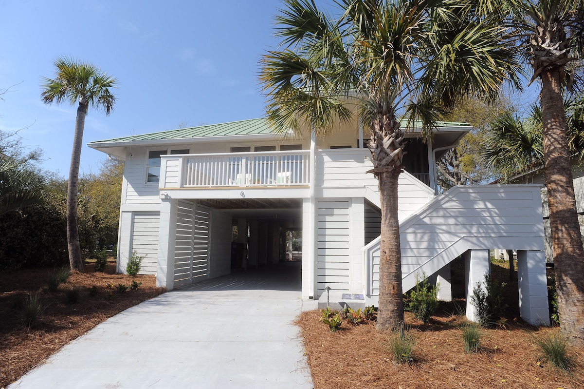 A two-story beach home features a light-colored exterior with a green metal roof. Palm trees are situated in the front yard, and a driveway leads to a covered carport beneath the home. A set of wooden stairs is visible on the side, providing access to the upper level.
