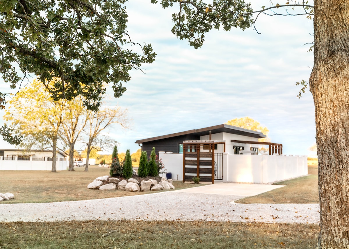 A modern villa is set against a clear sky, surrounded by green grass and trees. The sleek exterior features a combination of dark wood and white walls. A landscaped area with shrubs and rocks adds a touch of nature, enhancing the villa's inviting entry.