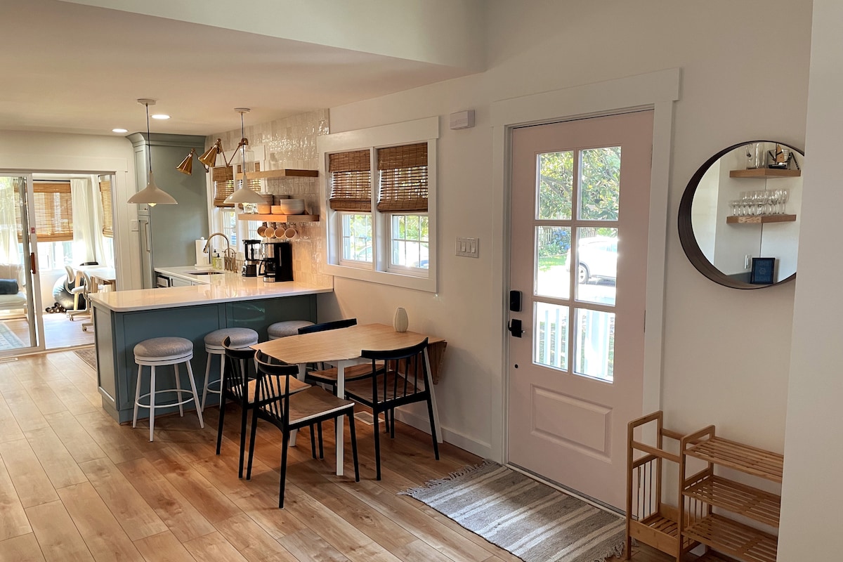 A light-filled kitchen and dining area features a spacious island with bar seating and modern fixtures. A dining table with black chairs sits on natural wood flooring, accompanied by a decorative mirror and a cozy entryway. Large glass doors lead to an outdoor space.