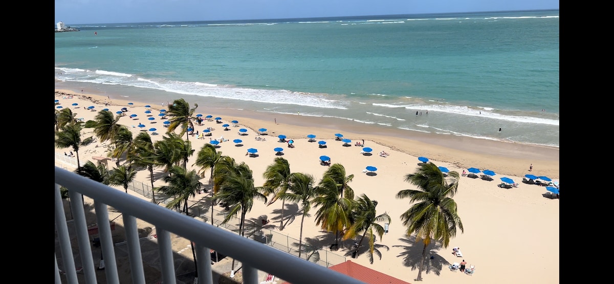 A panoramic view of a sandy beach stretches beneath a blue sky. Colorful umbrellas dot the shoreline, providing shade for beachgoers. Gentle waves lap at the shore, and palm trees sway in the breeze, adding to the coastal scenery.