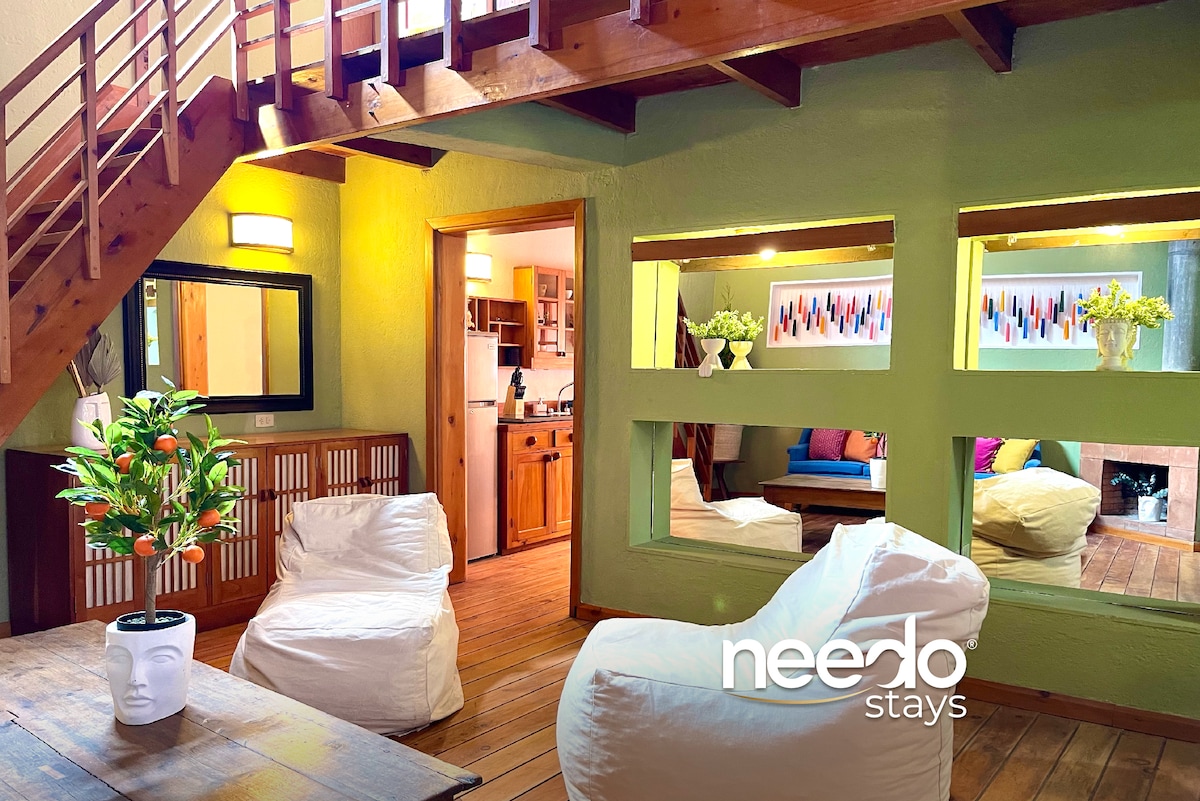 A cozy living area features light green walls complemented by wooden accents. Soft bean bag chairs are arranged around a rustic table. Openings provide views into other rooms, showcasing vibrant decor. A kitchen area is visible in the background, adding to the home's inviting layout.