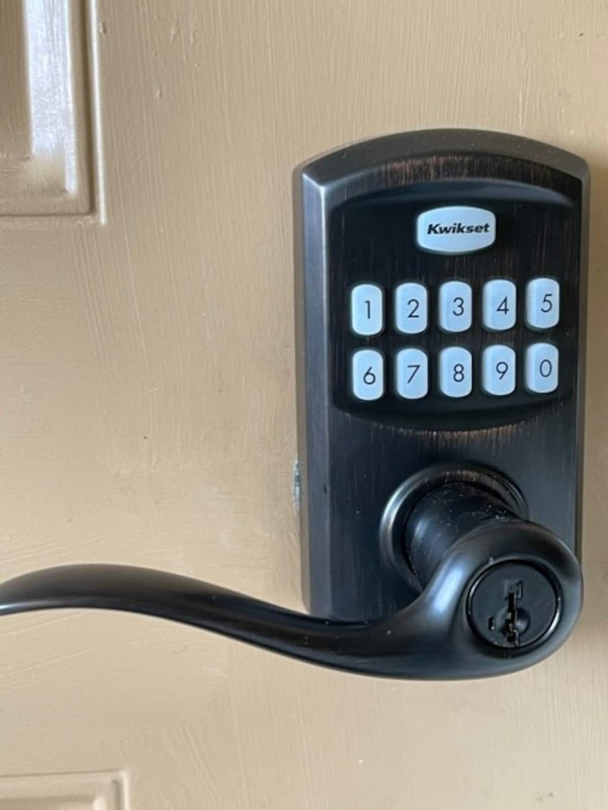 A keyless entry system is mounted on a brown door. The modern lock features a numeric keypad with large, easy-to-read numbers and a black handle with a keyed lock. The design combines security with convenience.