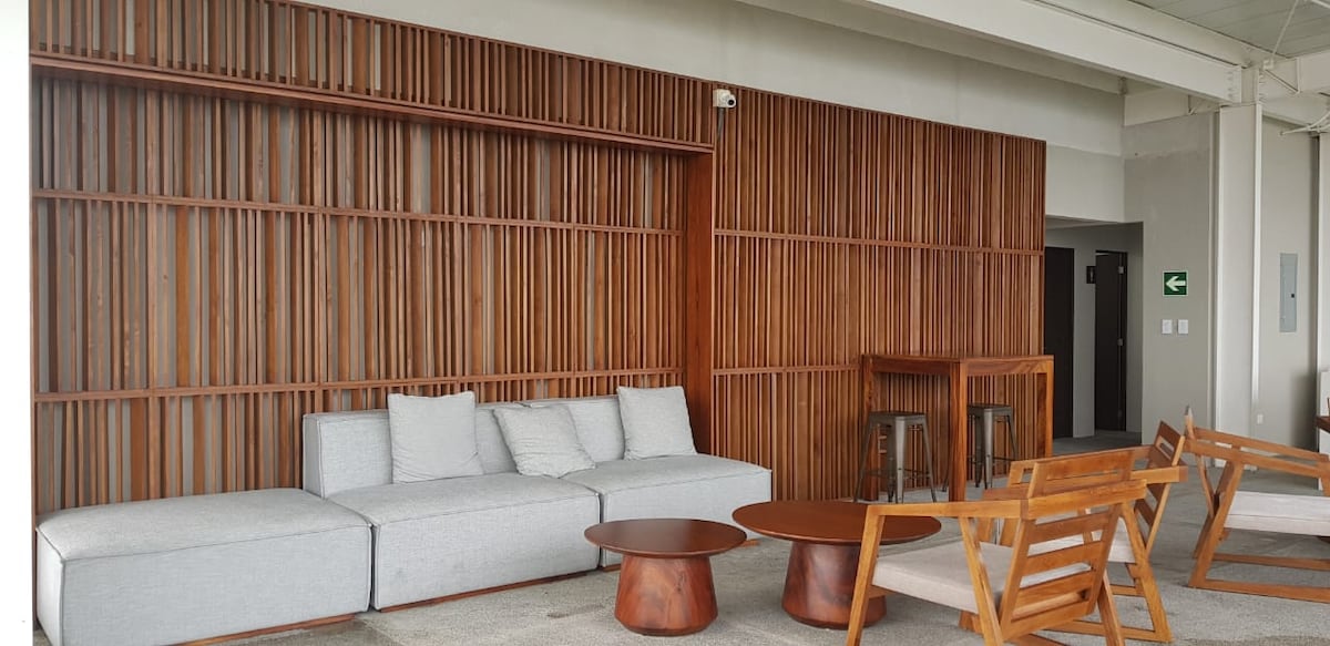 A contemporary lounge area features a wooden accent wall, complemented by a light gray sectional sofa. A round wooden coffee table and wooden chairs are arranged to create a comfortable seating arrangement. Natural light enhances the modern ambiance, inviting relaxation.