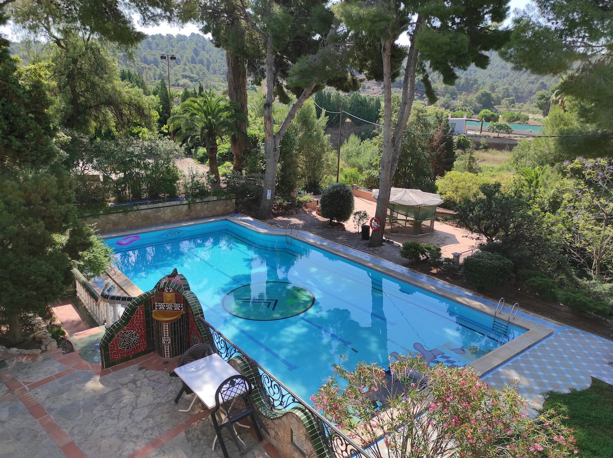 A serene outdoor space features a large swimming pool surrounded by lush greenery and various trees. Sun loungers are positioned nearby, and a gazebo offers shaded seating. The vibrant blue water reflects the clear sky and nearby plants, creating a refreshing ambiance.