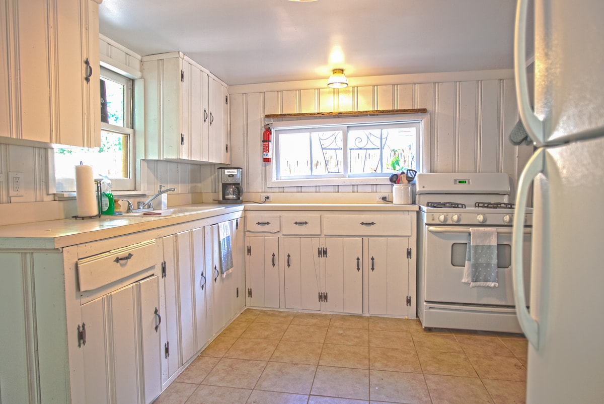 A bright kitchen features white cabinetry and tiled flooring, complemented by a window that allows natural light to fill the space. Essential appliances, including a stove and coffee maker, are visible, along with ample counter space for meal preparation.