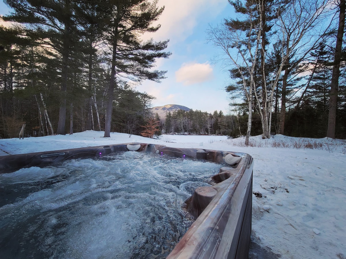 The outdoor hot tub is surrounded by snow-covered ground and tall trees, offering a serene view of the mountains in the distance. The bubbling water invites relaxation, with clear blue skies and soft clouds overhead.