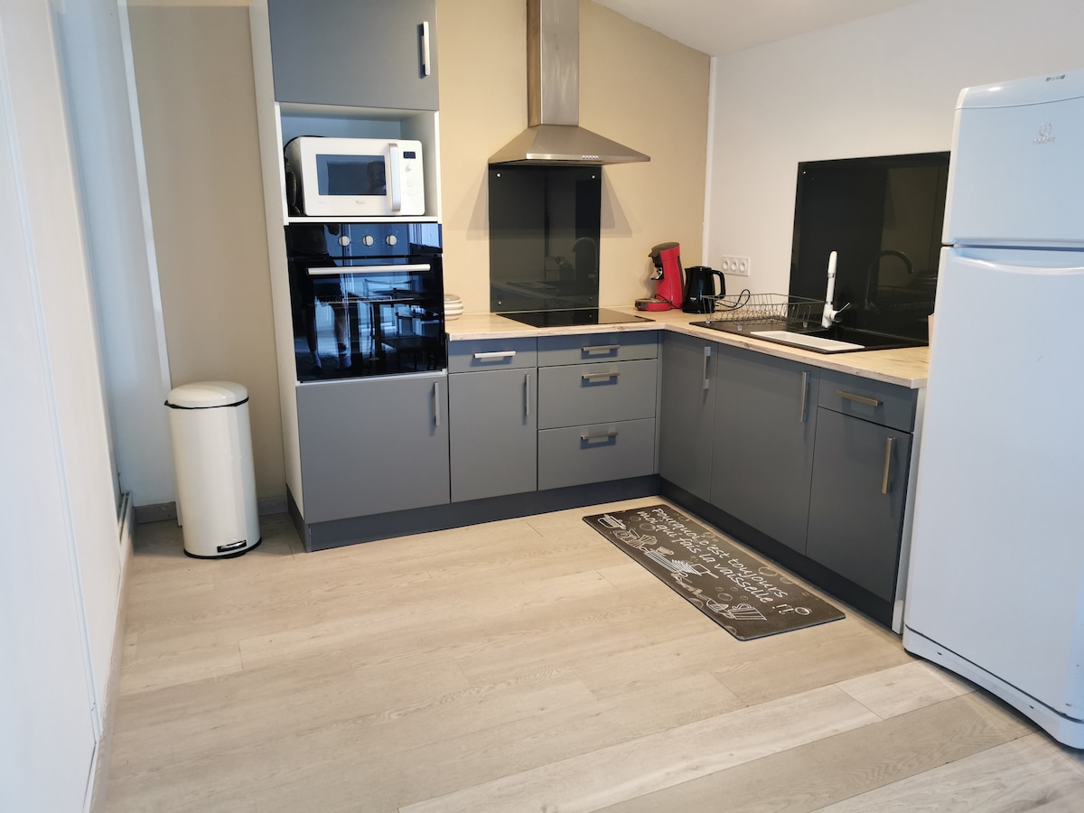 The kitchen area showcases modern cabinetry with grey finishes. Appliances include a microwave, coffee maker, and black oven. A white refrigerator stands against the wall. The space features wood-like flooring and a mat on the floor under the sink, providing a functional and clean appearance.