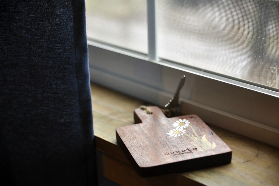 A wooden cutting board featuring a floral design is positioned on a light-colored window sill. Light filters through a sheer curtain, creating a soft ambiance in the space. The window provides a view of the outdoors, enhancing the inviting atmosphere.
