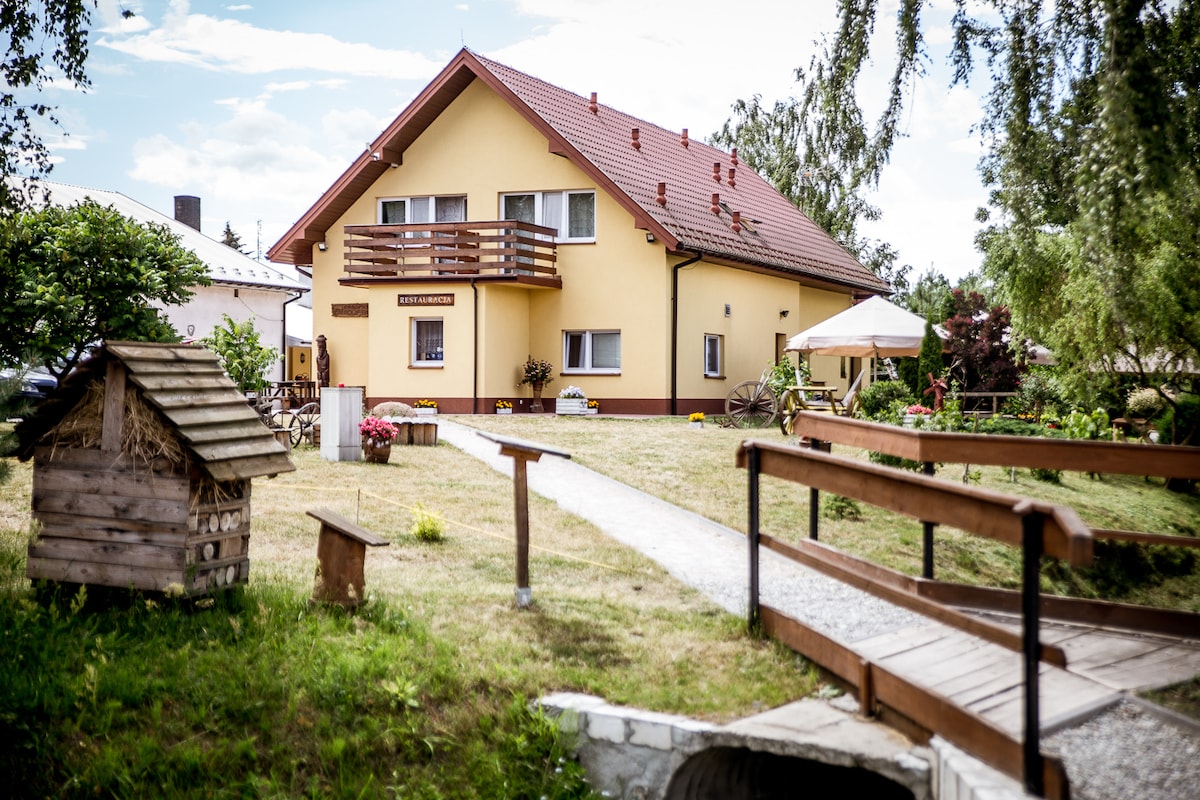 A welcoming rural property is shown, featuring a yellow facade and a sloped red roof. A garden area with pathways is visible, complemented by wooden structures and a sitting area. Lush greenery surrounds the building, enhancing the peaceful outdoor atmosphere.