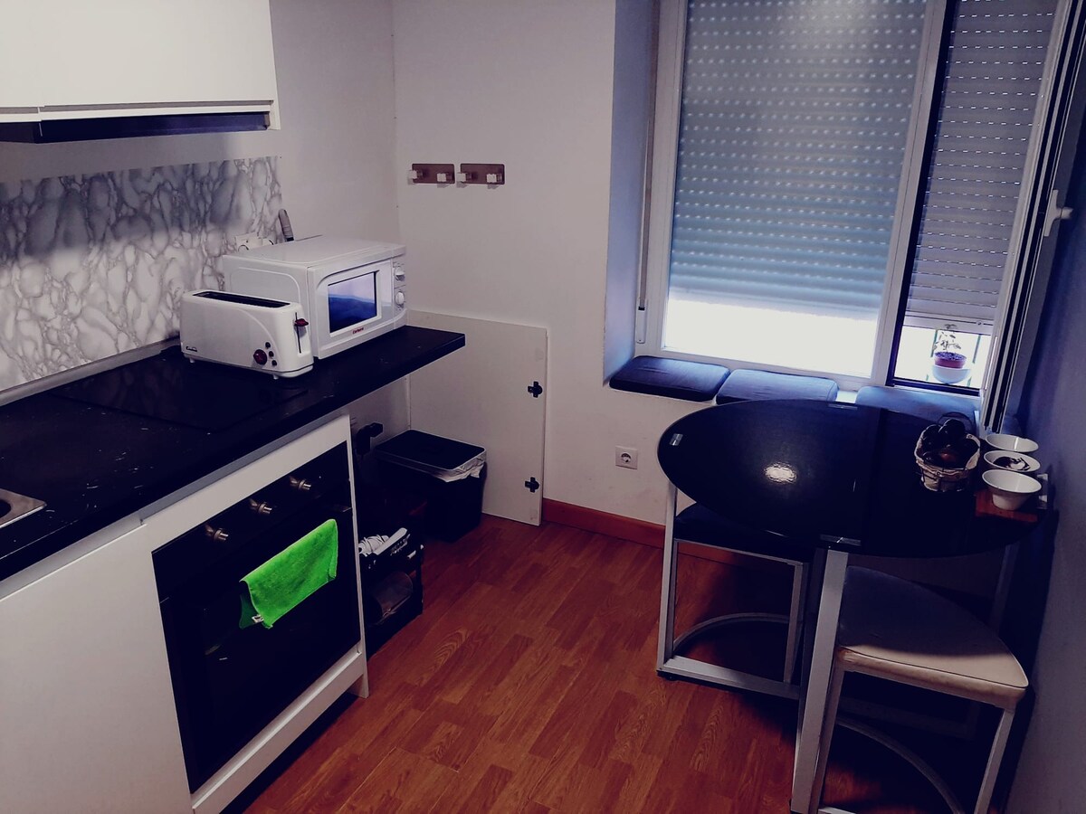 The kitchen features modern appliances, including a microwave and oven, set against a marble-patterned backsplash. A small round table is positioned near a window, with two white chairs. Soft light is filtered through the window's shades, creating a functional cooking space.