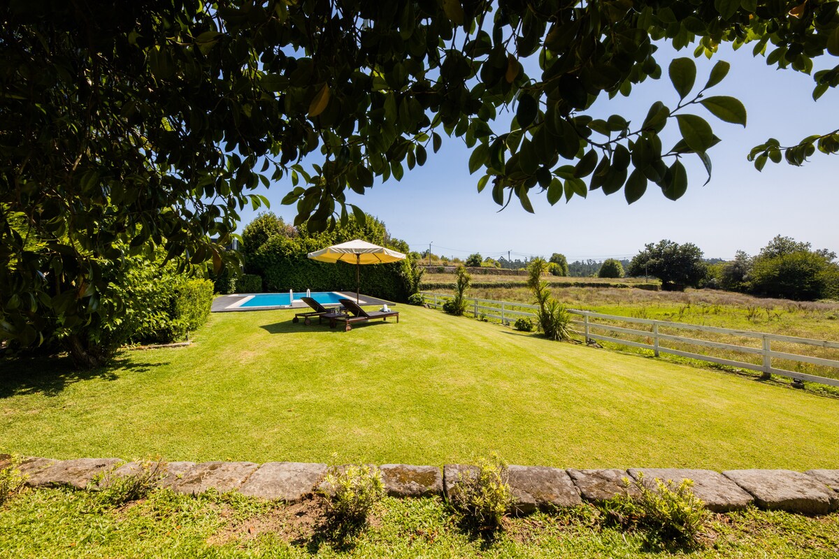 A well-maintained lawn extends towards a serene outdoor pool, accompanied by lounge chairs and a sun umbrella. The space is shaded by leafy branches, providing a tranquil setting surrounded by gardens and distant views of lush greenery and trees.