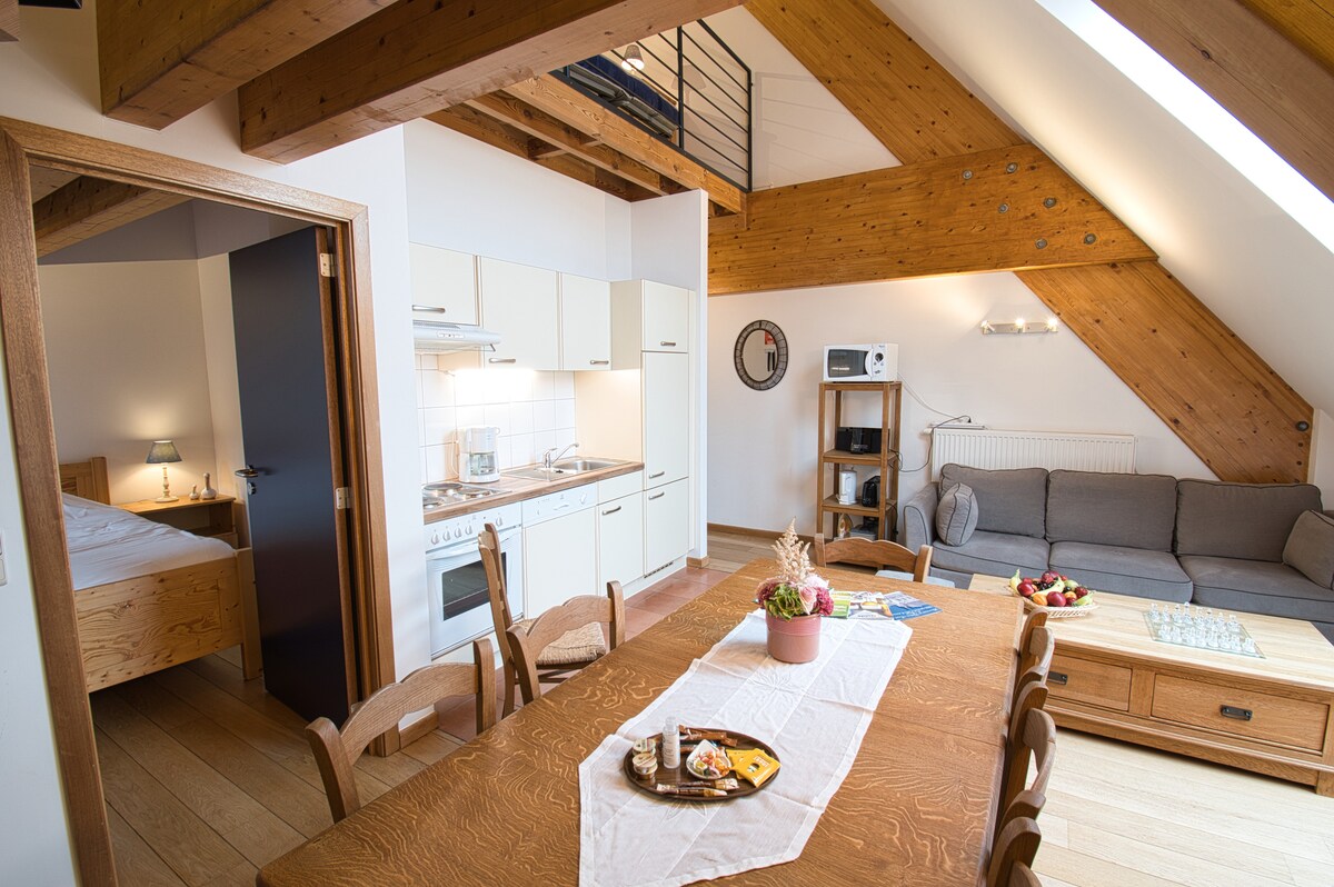 An open-plan living area displays wooden beams and natural light from large windows. A dining table sits centrally, surrounded by wooden chairs. The kitchenette is equipped with modern appliances. A cozy seating area features a gray sofa, adding to the welcoming environment.