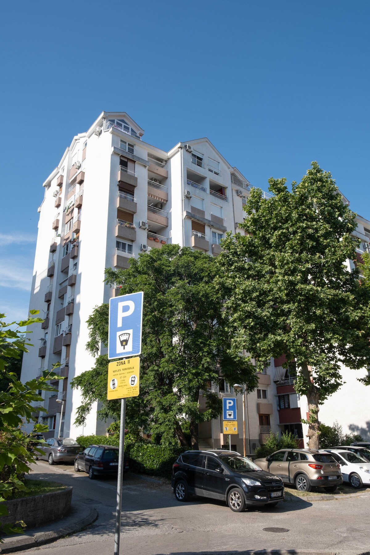 A multi-story building is prominently displayed, featuring a clean white façade and several balconies. Lush green trees partially frame the scene, and a parking area with clear signage is visible in the foreground, emphasizing convenience for guests.