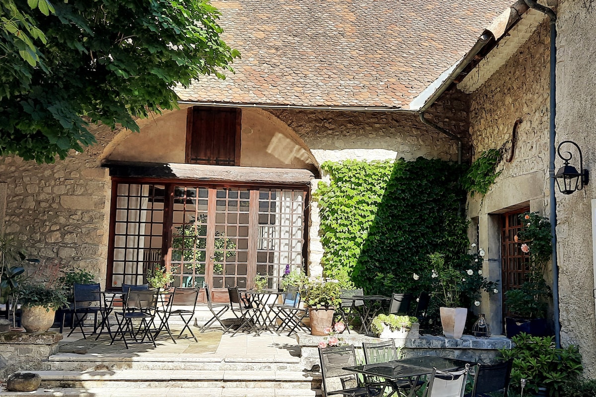 A charming outdoor area is presented, featuring a stone terrace lined with potted plants and vibrant greenery. Several black metal chairs surround small tables, providing seating space. Large glass doors lead into the indoor area, inviting light and creating a seamless flow between the exterior and interior.