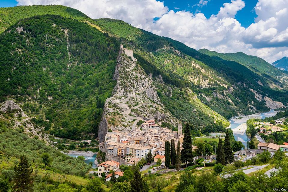 The image captures a panoramic view of a medieval village nestled between lush green mountains, featuring a small cluster of historic buildings. A river flows nearby, and a medieval castle sits atop a rocky outcrop, creating a scenic contrast against the natural landscape.