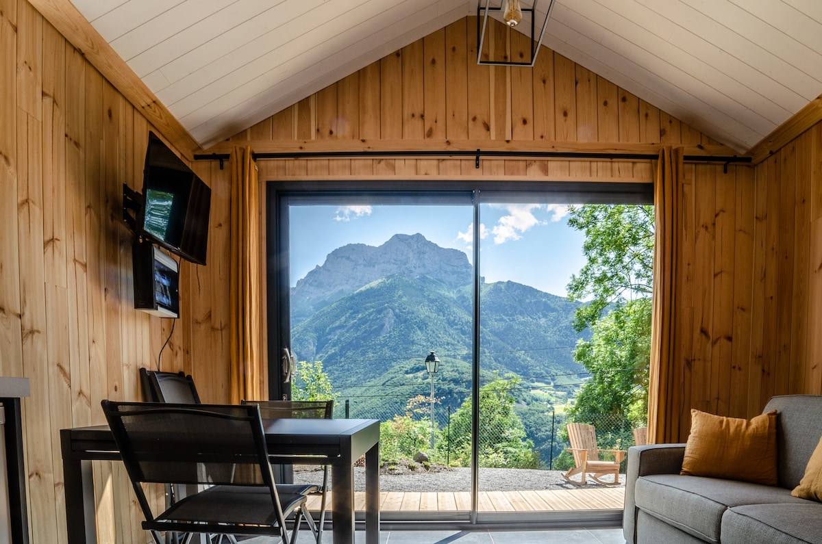 A wooden interior features large glass doors that offer a panoramic view of surrounding mountains. A dining table is positioned beside the doors, with two chairs and a sofa creating a comfortable seating area. Warm yellow curtains frame the view, adding to the natural ambiance.