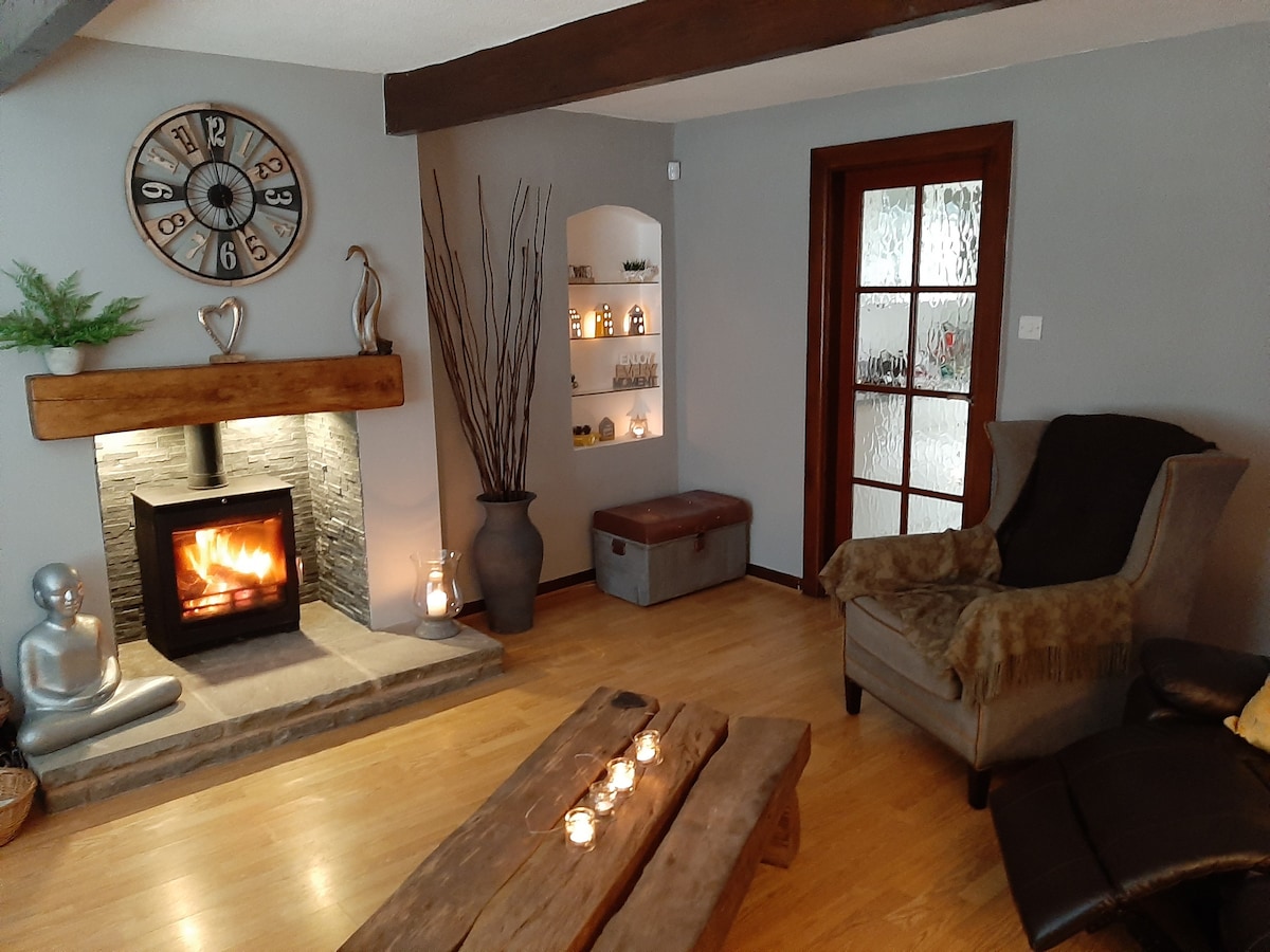 A welcoming lounge area features a stone fireplace with a warm flame, complemented by wooden beams on the ceiling. Comfortable seating includes an armchair draped with a soft throw. A decorative coffee table is arranged with candles, while built-in shelves display various decor items.