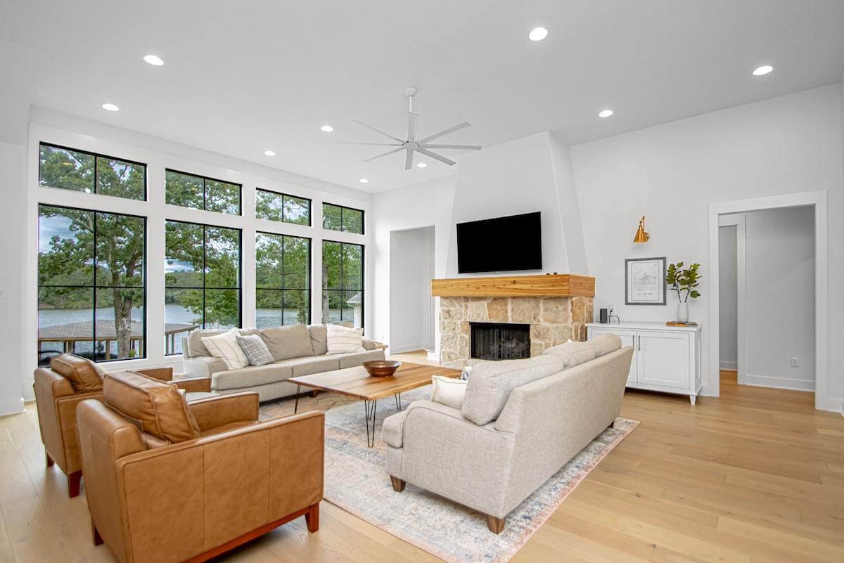 The living area is bright and spacious, featuring large windows that provide views of the surrounding nature and lake. A stone fireplace is centered against a light wall, with a large flat-screen TV mounted above it. Various seating options, including leather and fabric sofas, complement a central coffee table.