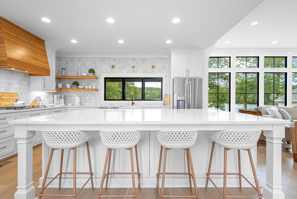 The modern kitchen features a large island with seating for four, illuminated by recessed lighting. Natural wood accents are complemented by white cabinetry and a stylish marble backsplash. A view of the lake is visible through the expansive windows, enhancing the inviting atmosphere.