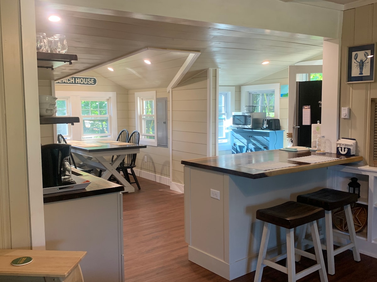A bright and open kitchen area is showcased, featuring wooden countertops and bar stools. Large windows allow natural light to fill the space, complementing the neutral wall tones. A dining table with chairs is visible in the background, emphasizing a welcoming atmosphere.