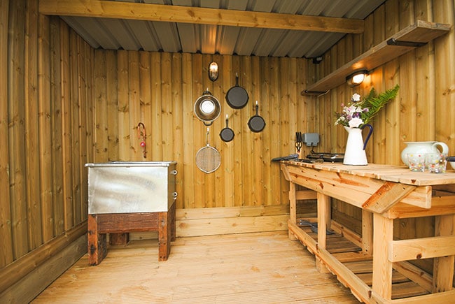 An outdoor kitchenette features wooden walls with a polished wood floor. A stainless steel sink is fitted to the left, with hanging cooking utensils and a shelf above. A rustic wooden table holds kitchen equipment, providing an organized and functional space for meal preparation.