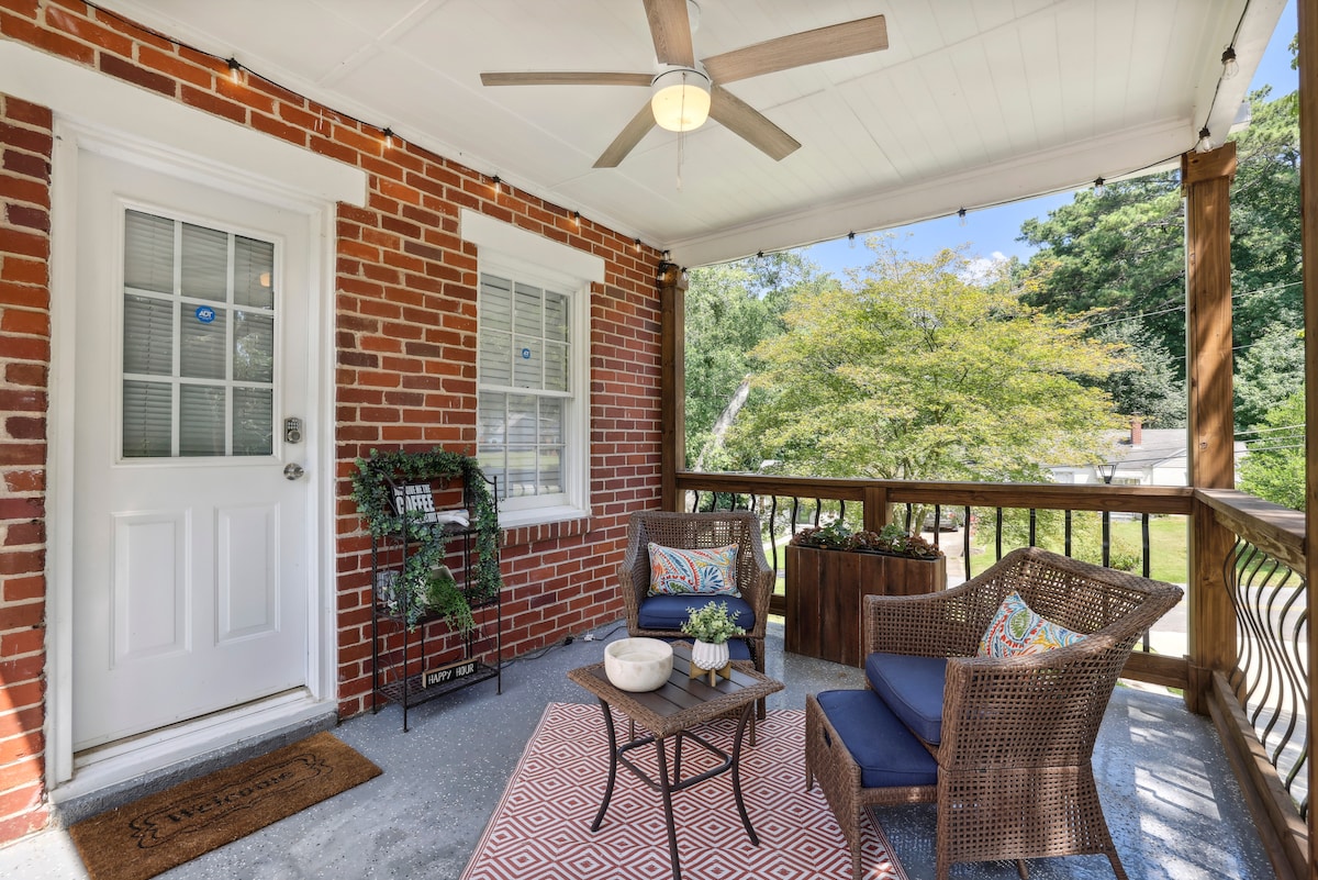 A welcoming porch area features two comfortable chairs arranged around a small table. A gentle ceiling fan provides cooling relief, while greenery adds a touch of nature. The brick facade complements the inviting outdoor space, perfect for relaxing moments.