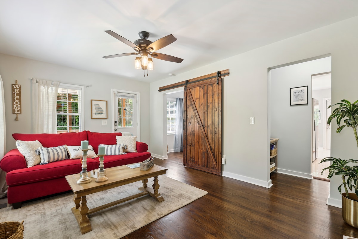 A bright living area features a vibrant red sofa adorned with decorative pillows. A wooden coffee table sits at the center on a light area rug. Natural light filters through windows, enhancing the inviting atmosphere. A rustic sliding barn door adds character to the space.