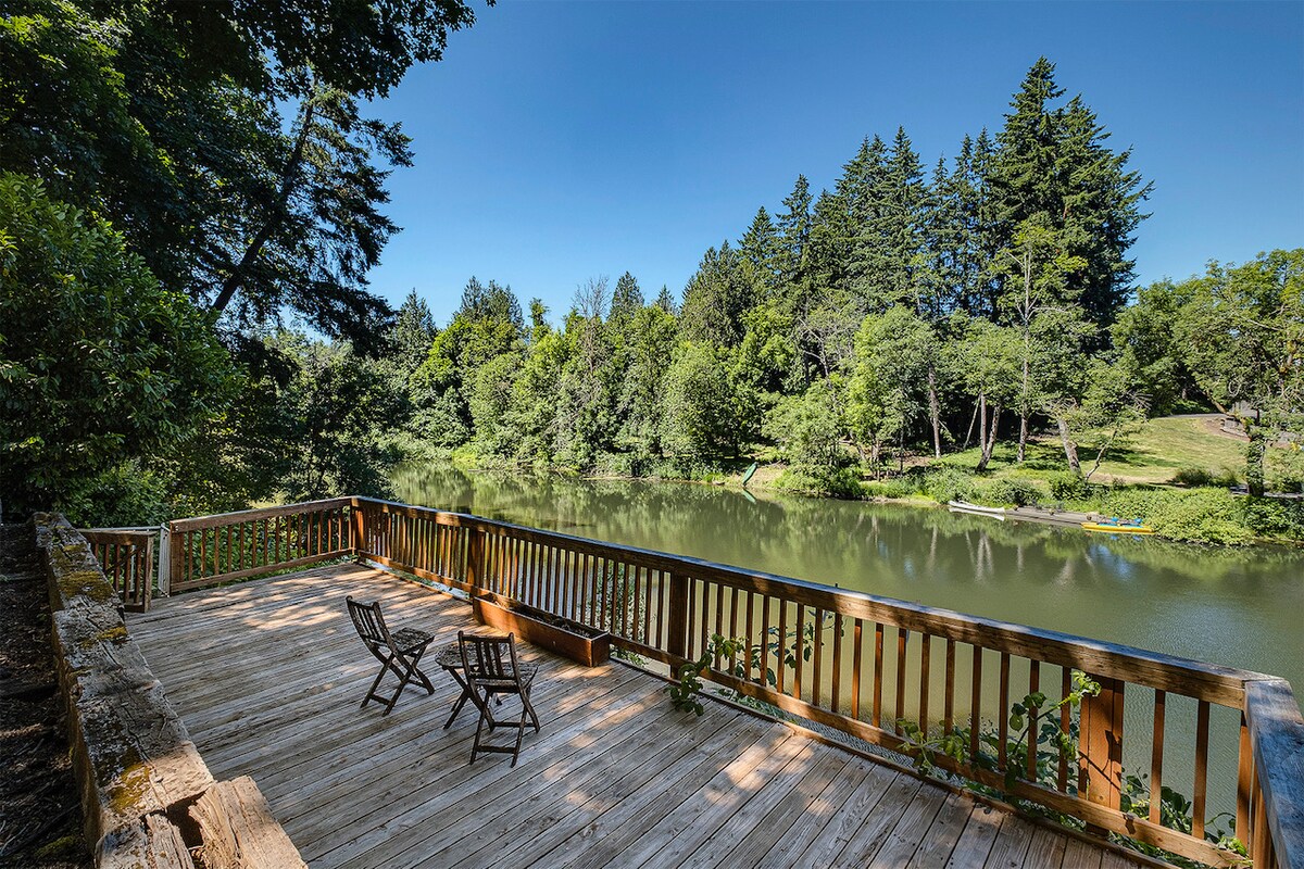 A wooden deck overlooks a peaceful river surrounded by lush greenery and tall trees. Two chairs are positioned on the deck, inviting guests to relax and enjoy the serene water views.