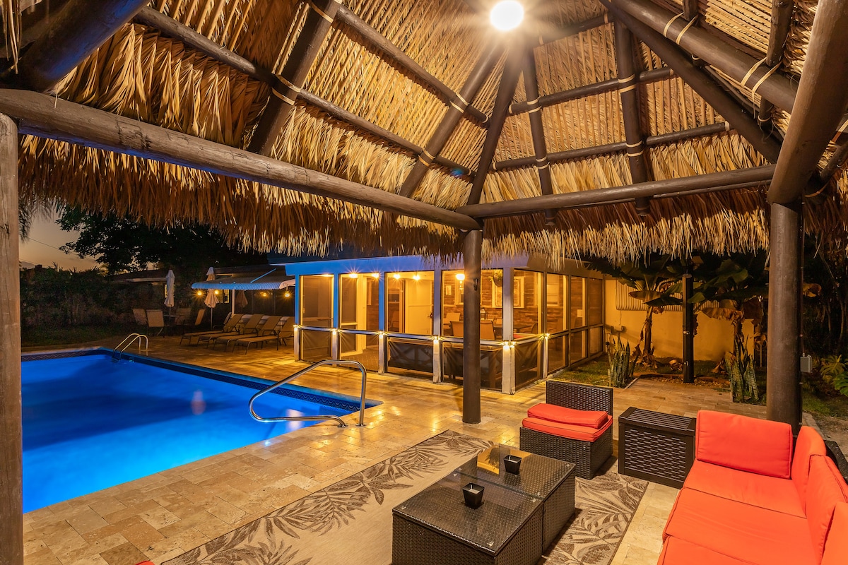 An outdoor Tiki Hut is framed by a thatched roof, providing shade over the seating area. An inviting heated pool with a bright blue surface is visible in the background, illuminated by soft lighting. The space features modern outdoor furniture and surrounded by lush tropical landscaping.